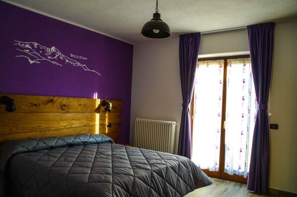 Une chambre à coucher avec un grand lit, des murs violets, une tête de lit en bois, un radiateur et une fenêtre ornée de rideaux violets.