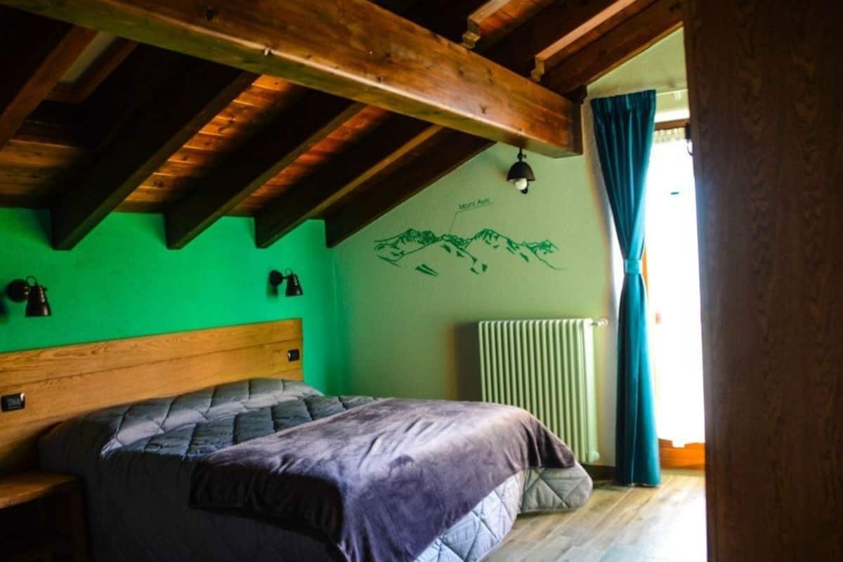 A bedroom with a wooden ceiling, a bed with a headboard, a green wall with a mountain drawing, a radiator, and a window with curtains.