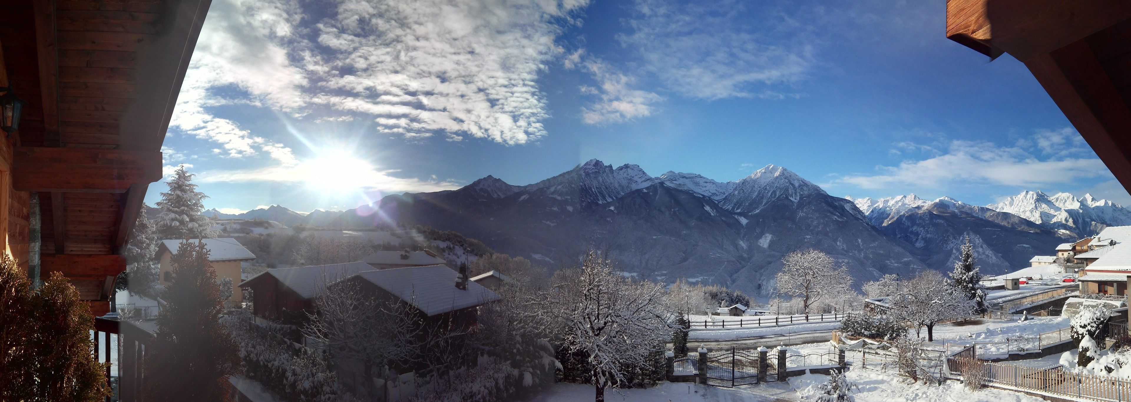 Blick auf Berge mit schneebedeckten Bäumen und Gebäuden, bewölkter Himmel und helle Sonne.