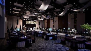 A spacious banquet hall with tables set for events, draped curtains, and a stage with floral arrangements.