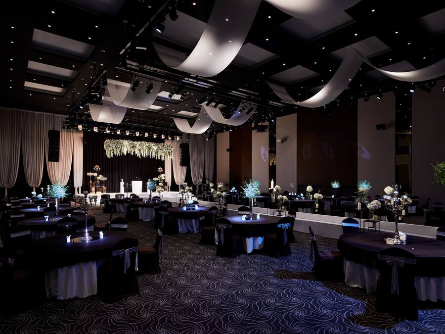 A spacious banquet hall with tables set for events, draped curtains, and a stage with floral arrangements.