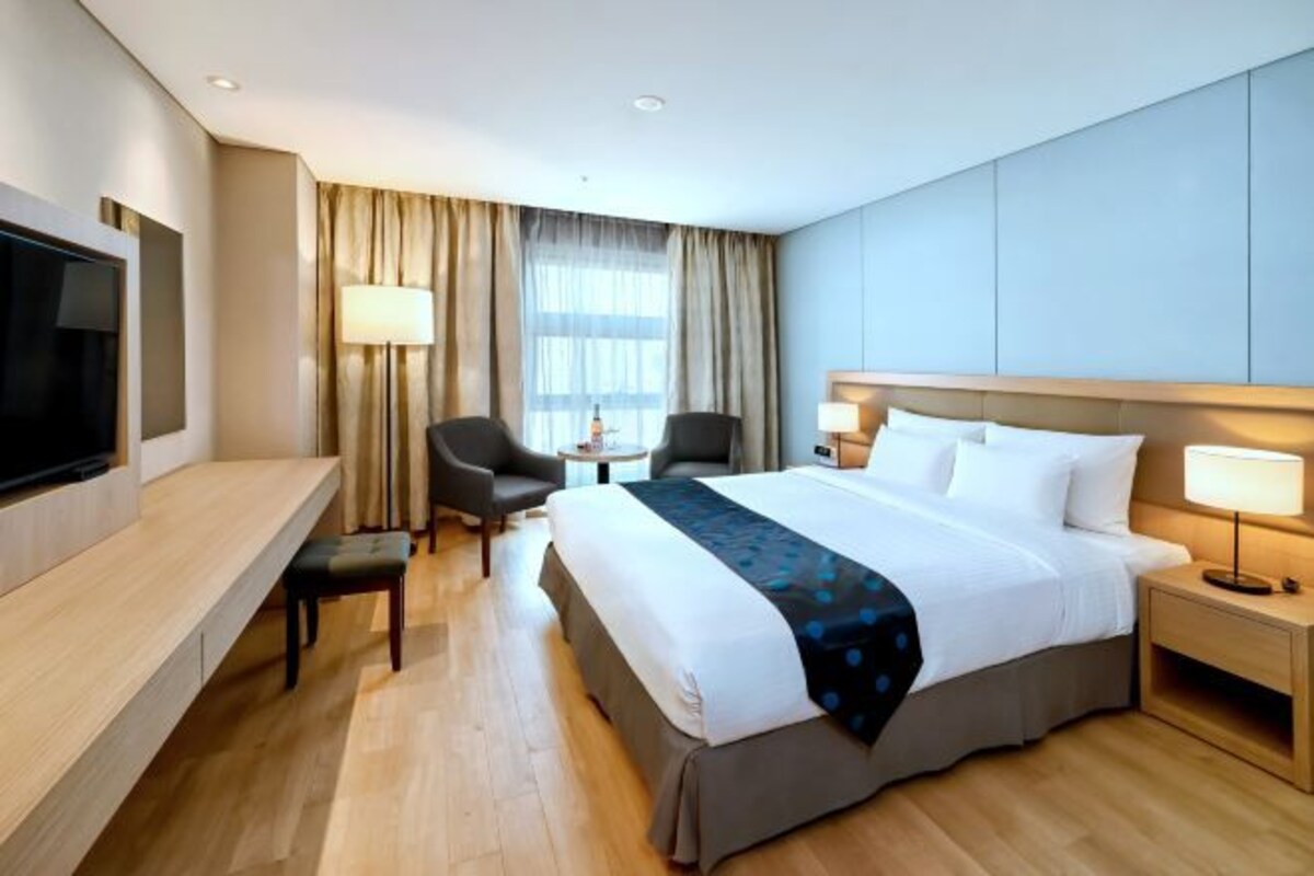 Habitación de hotel con una cama grande, dos mesitas de noche con lámparas, un televisor de pantalla plana en la pared, un escritorio pequeño con una silla y una ventana con cortinas.
