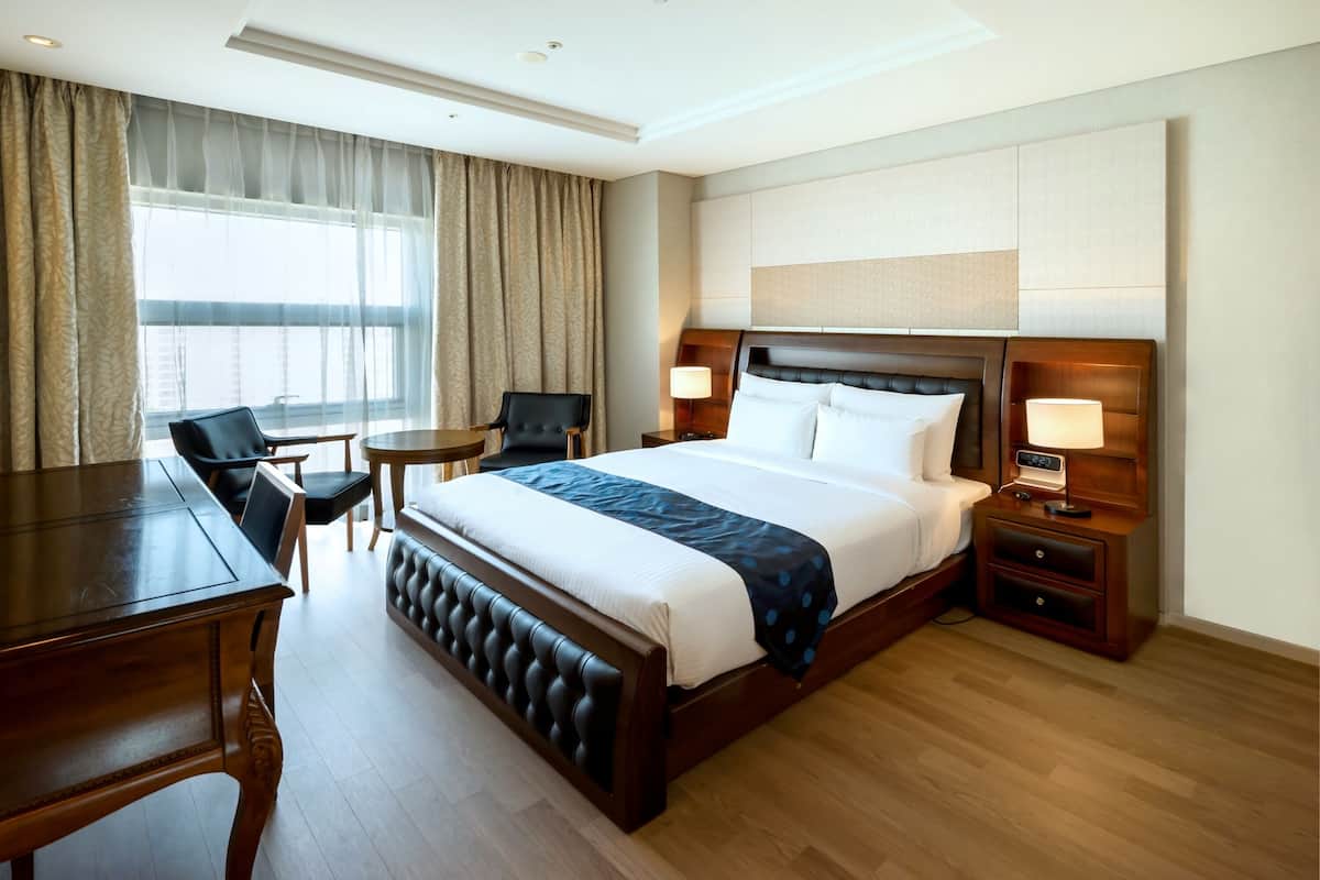 A hotel room with a large bed, a desk, a chair, a small table, and a nightstand with a lamp.