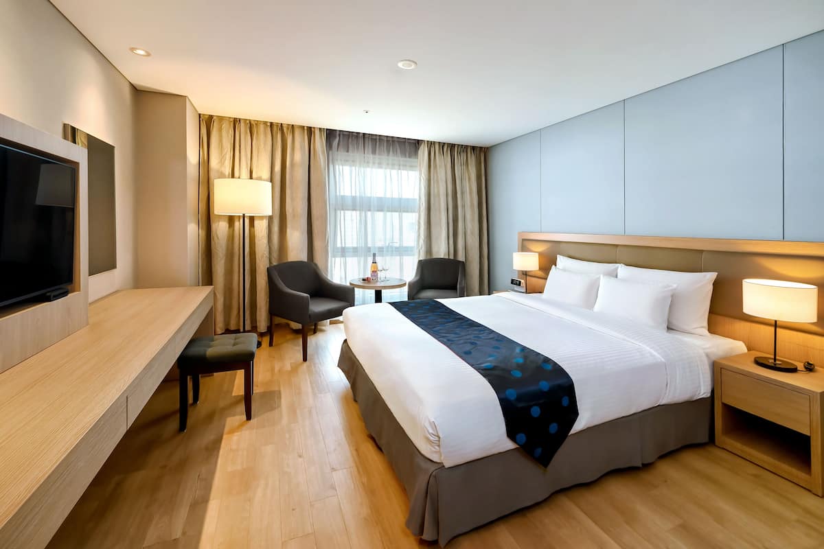 A modern hotel room with a large bed, bedside lamps, a desk with a flat-screen TV, and a seating area with two chairs.