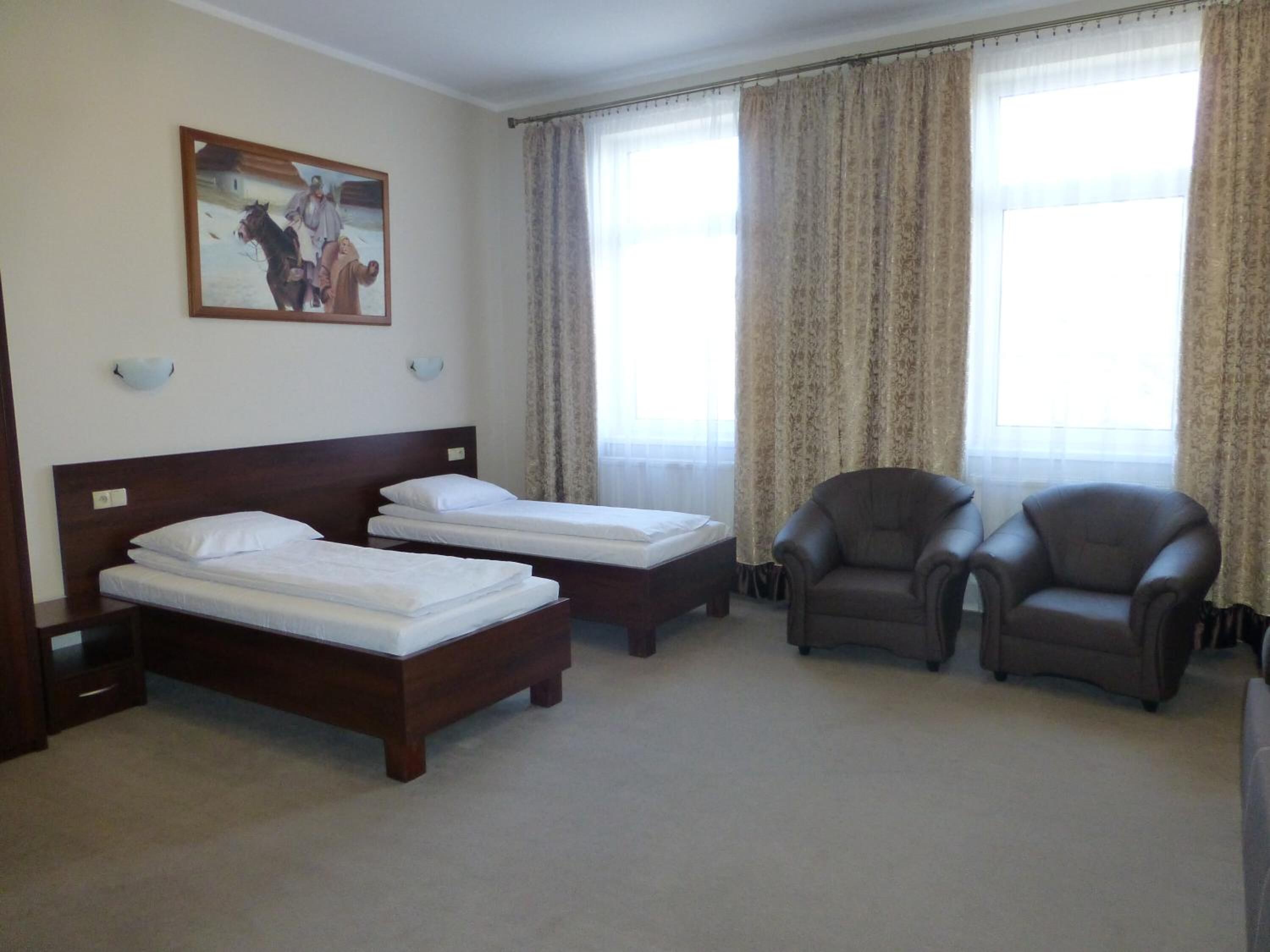 A hotel room with two single beds, a wooden headboard, a large window with curtains, and two leather armchairs.