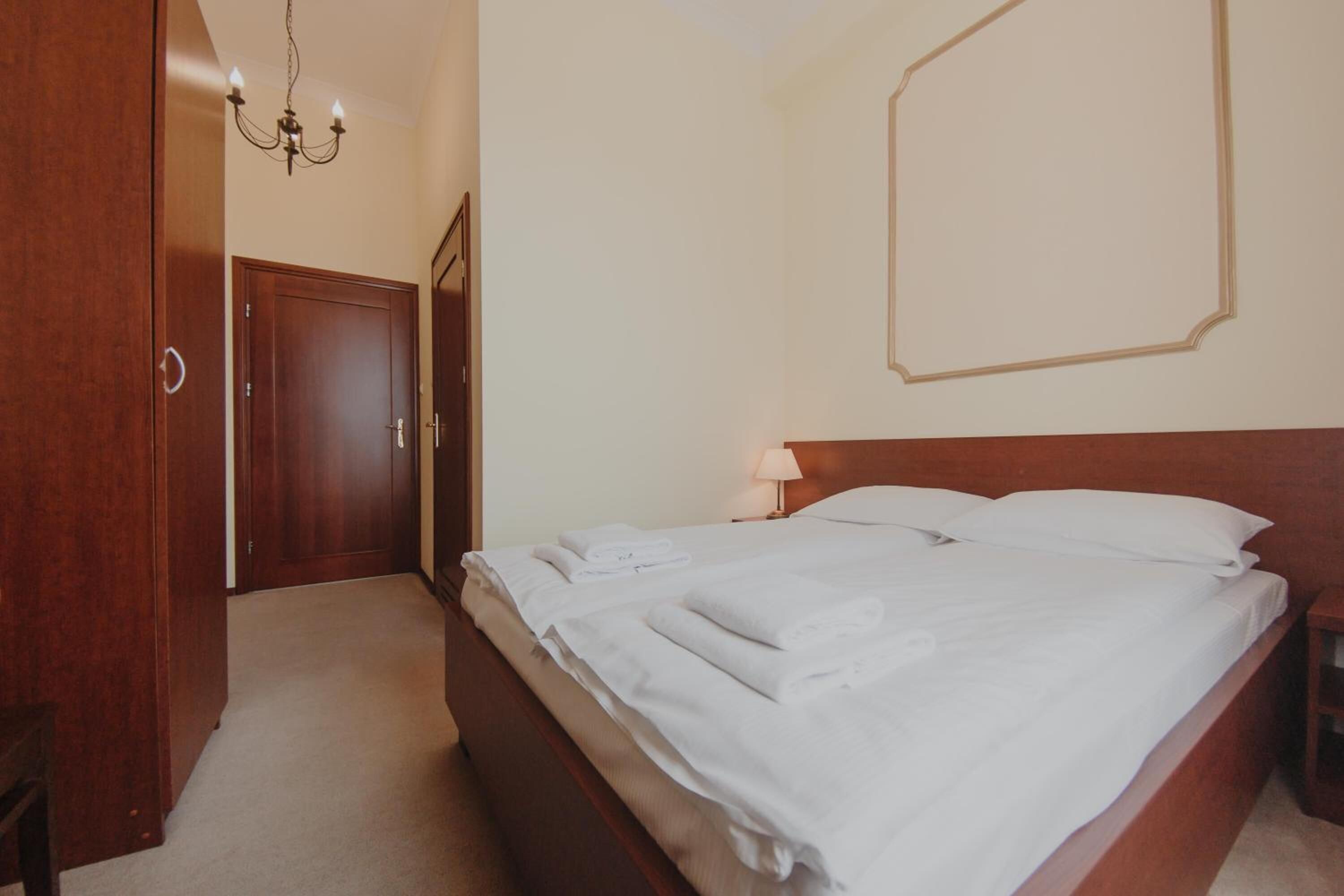 A bedroom with a wooden bed, white linens, a bedside table, a lamp, and a chandelier.
