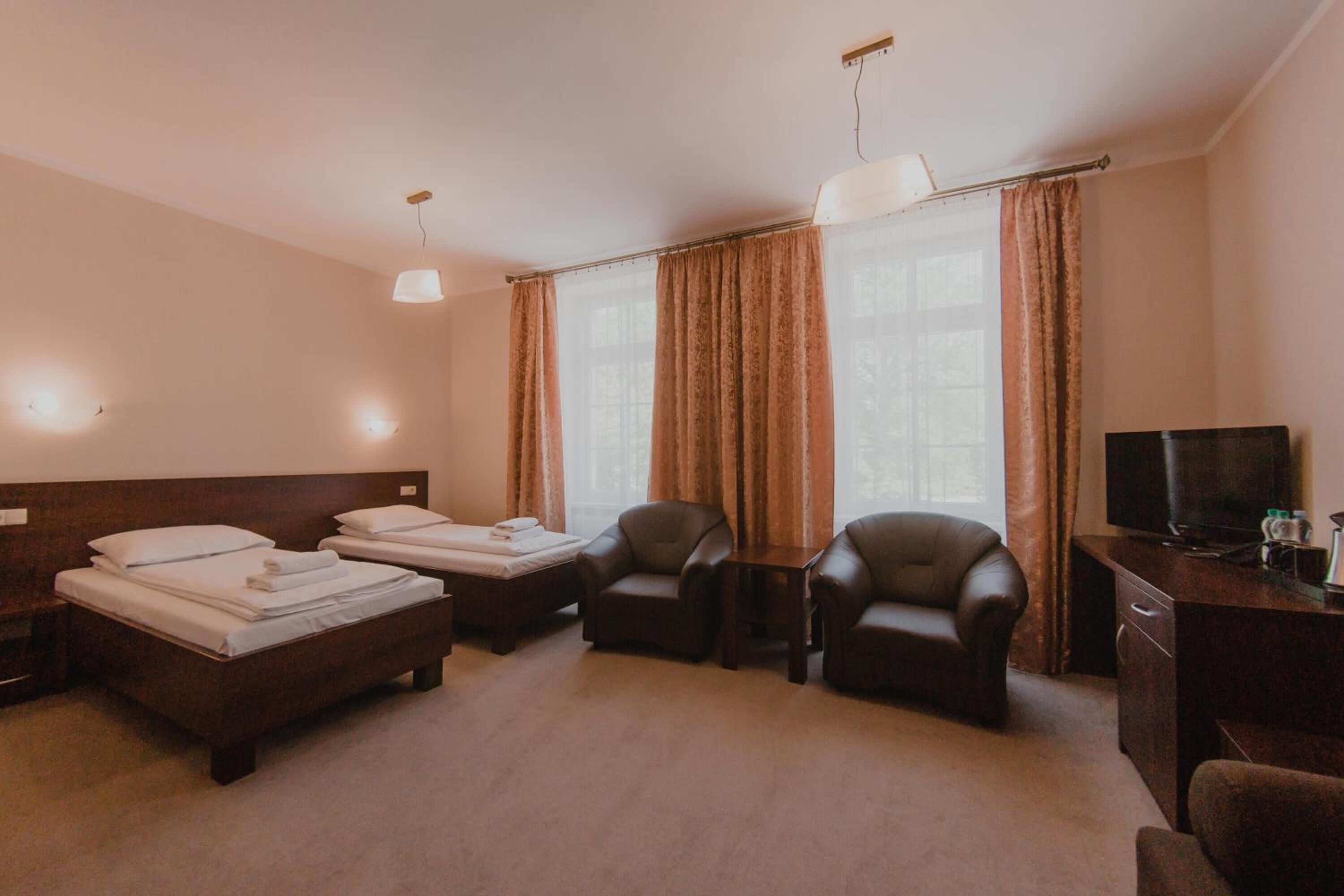 A hotel room with two beds, two leather armchairs, a desk with a television, and a large window with curtains.