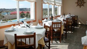 Un comedor con mesas dispuestas para una comida, amplias ventanas con cortinas, y vistas a edificios y al agua.