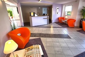 A modern hotel lobby with a reception desk, orange seating, and a tiled floor.