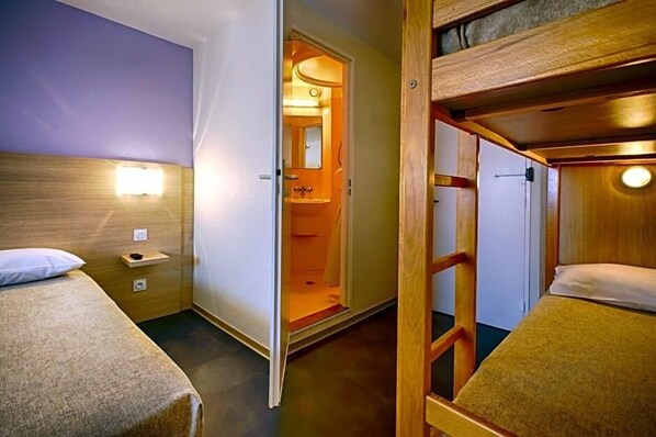 A compact room with bunk beds, a bedside table, a nightstand, a wall-mounted light, and an open door leading to a bathroom.
