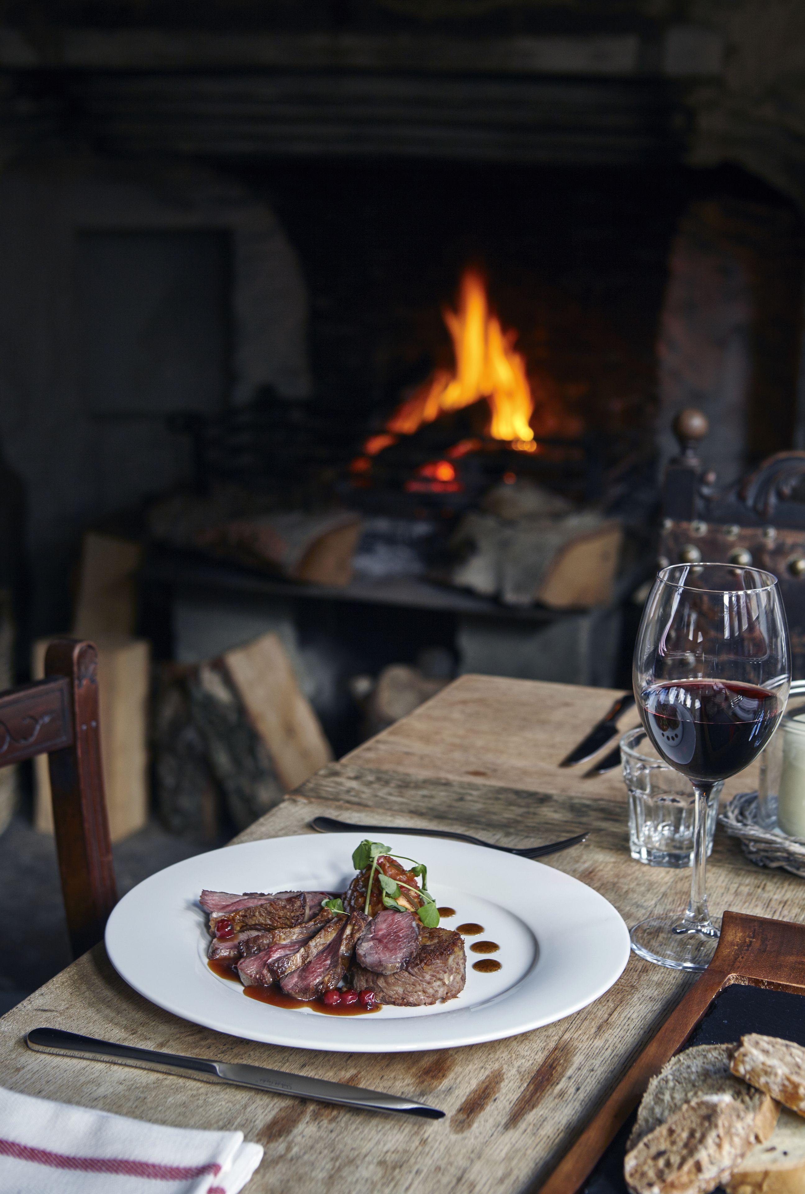 A dining table with a plate of food, a glass of red wine, and a fireplace in the background.
