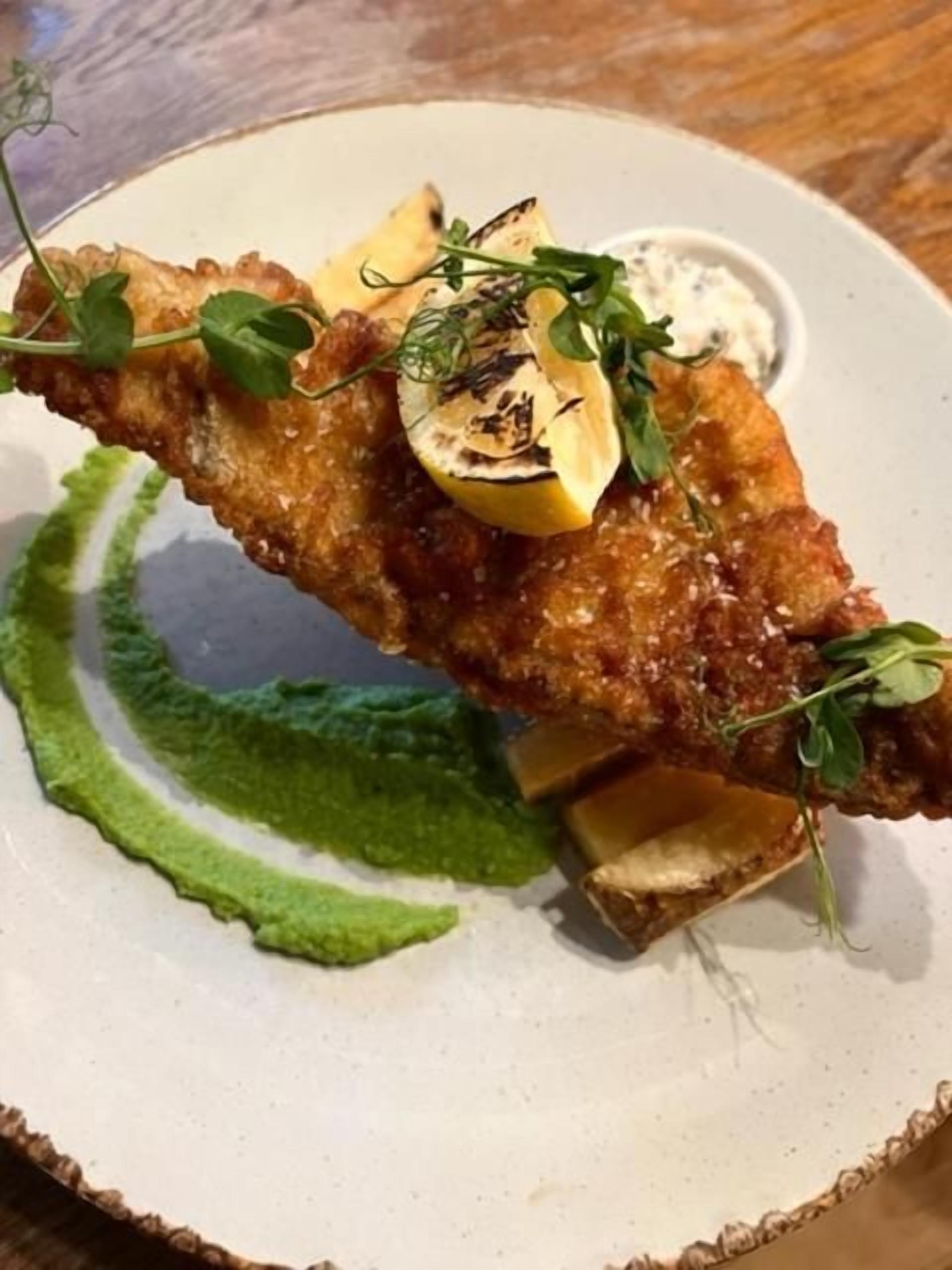 Een bord met een gepaneerd en gefrituurd gerecht, mogelijk een visfilet, versierd met citroen en kruiden, vergezeld van een groene saus en een klein schaaltje saus.