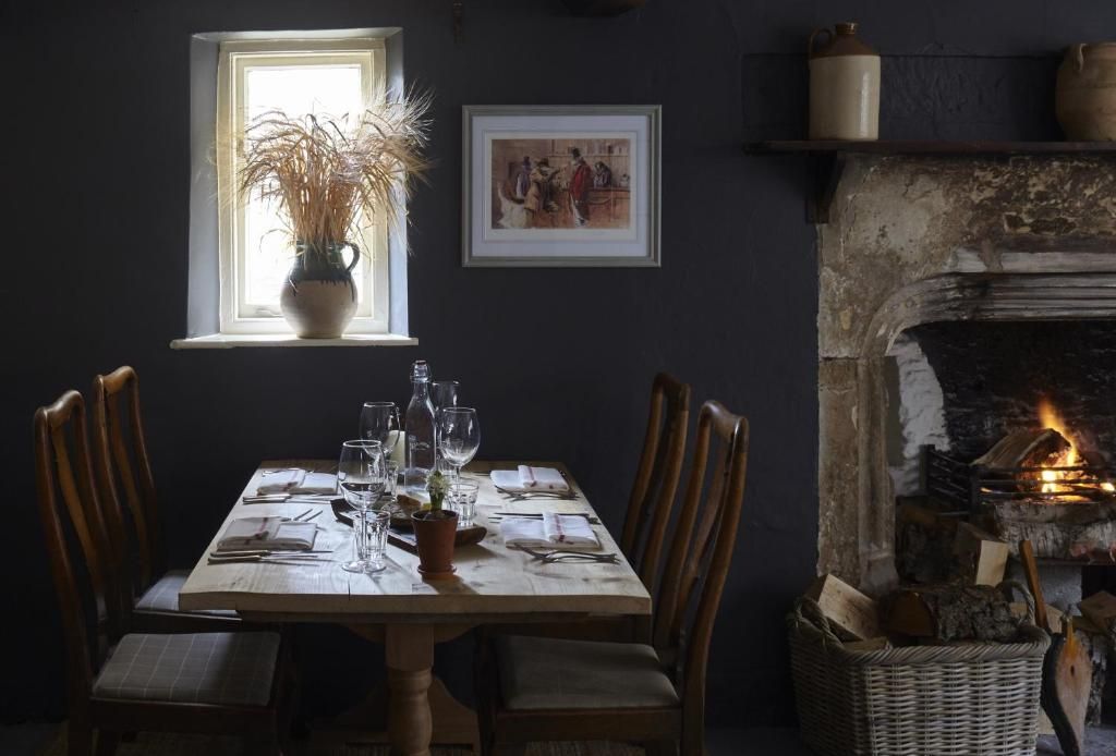 Una mesa de comedor dispuesta con sillas, un florero con plantas secas, una chimenea con leña y un cuadro en la pared.