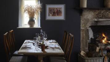 Una mesa de comedor dispuesta con sillas, un florero con plantas secas, una chimenea con leña y un cuadro en la pared.