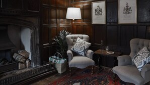 A traditional room with a fireplace, two armchairs, a table with a lamp, and framed coats of arms on the wall. - Sign of the Angel (Chippenham)