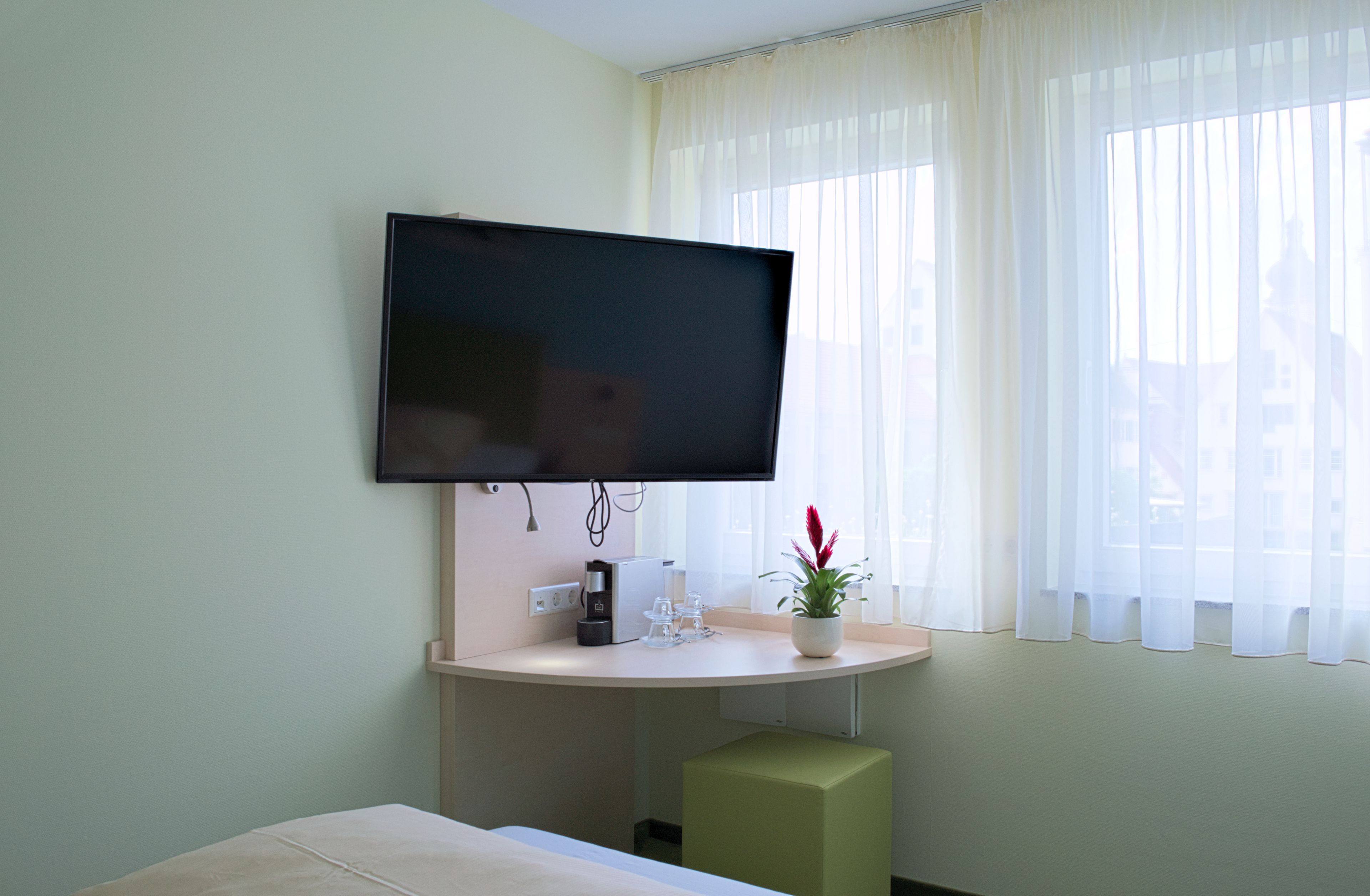 Habitación de hotel con televisor de pantalla plana en la pared, una mesita con cafetera, vasos y una planta en maceta.