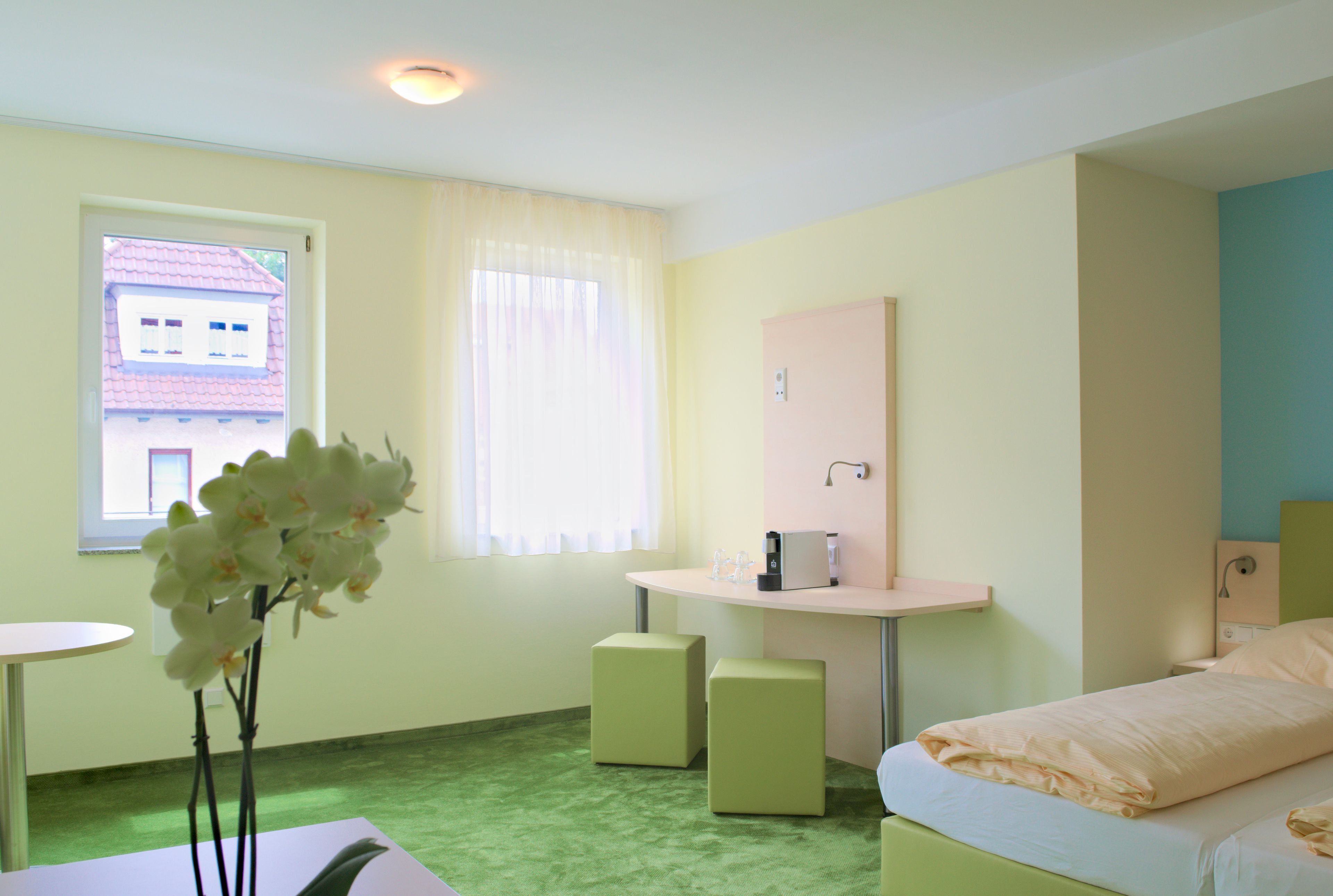 Una habitación de hotel con cama, mesita de noche, un escritorio pequeño con silla, ventana con cortinas y vista a un edificio.