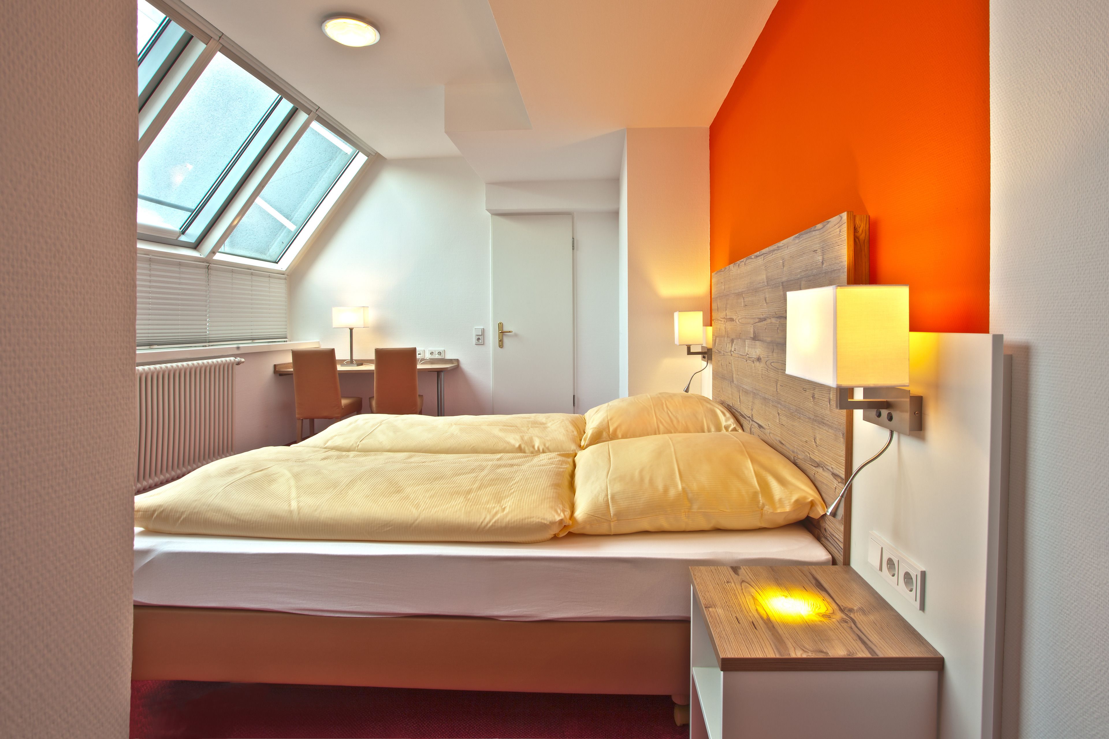 Een moderne slaapkamer met dakraam, een bed met geel beddengoed, een nachtkastje met lamp en een houten hoofdbord.