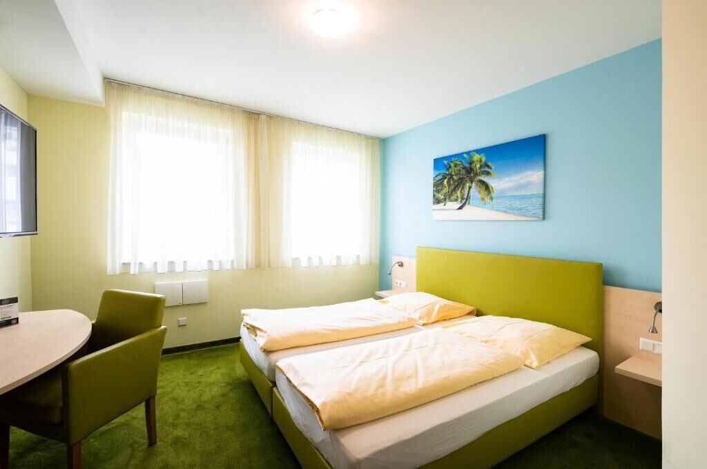 A hotel room with two single beds, a desk, a chair, a painting of a beach, and a window with curtains.