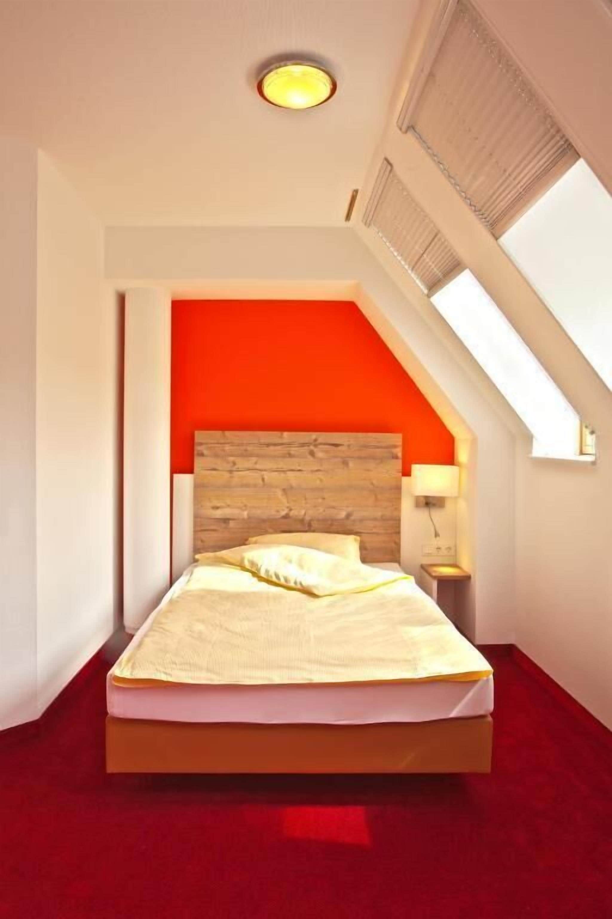 Une chambre avec un plafond en pente, une tête de lit en bois, un mur d’accent rouge, un lit avec une literie blanche et une table de chevet avec une lampe.