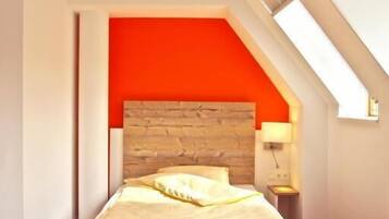 Une chambre avec un plafond en pente, une tête de lit en bois, un mur d’accent rouge, un lit avec une literie blanche et une table de chevet avec une lampe.