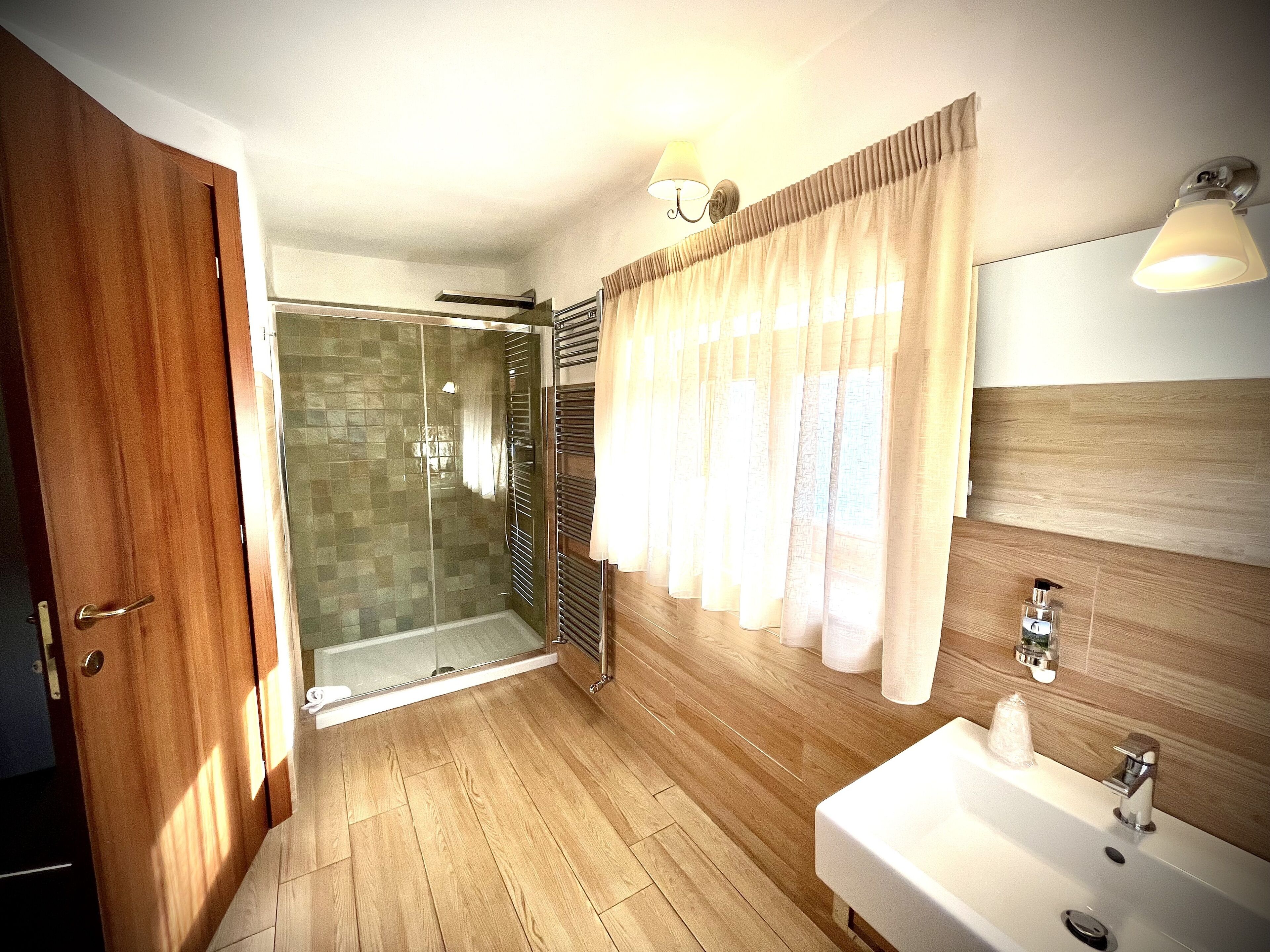 A bathroom with a wooden floor, a glass shower enclosure, a white sink, and a mirror with a wooden panel above it.