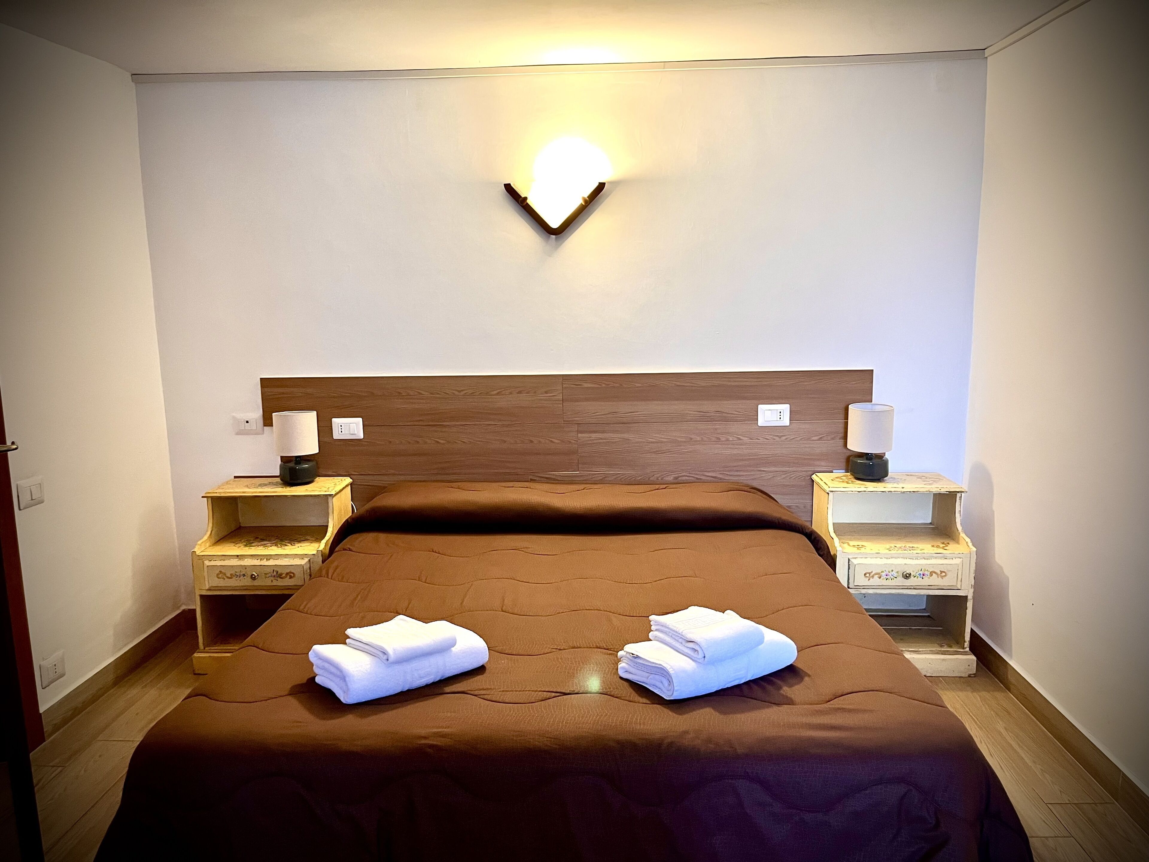 A hotel room with a wooden headboard, two bedside tables with lamps, a brown bedspread, and two folded white towels on the bed.