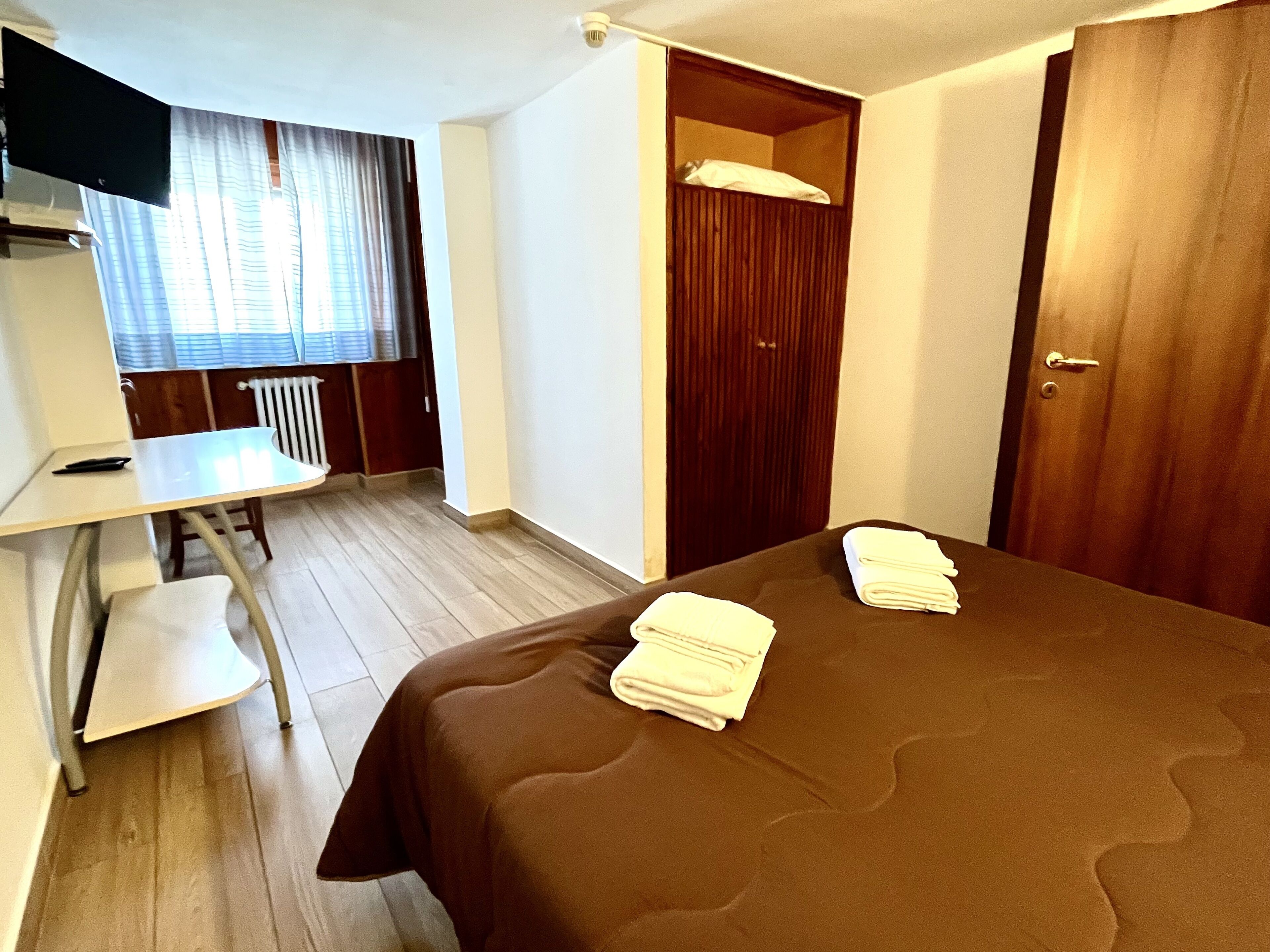 Une chambre d’hôtel avec un lit, un bureau, une chaise, une armoire et une fenêtre avec des rideaux.