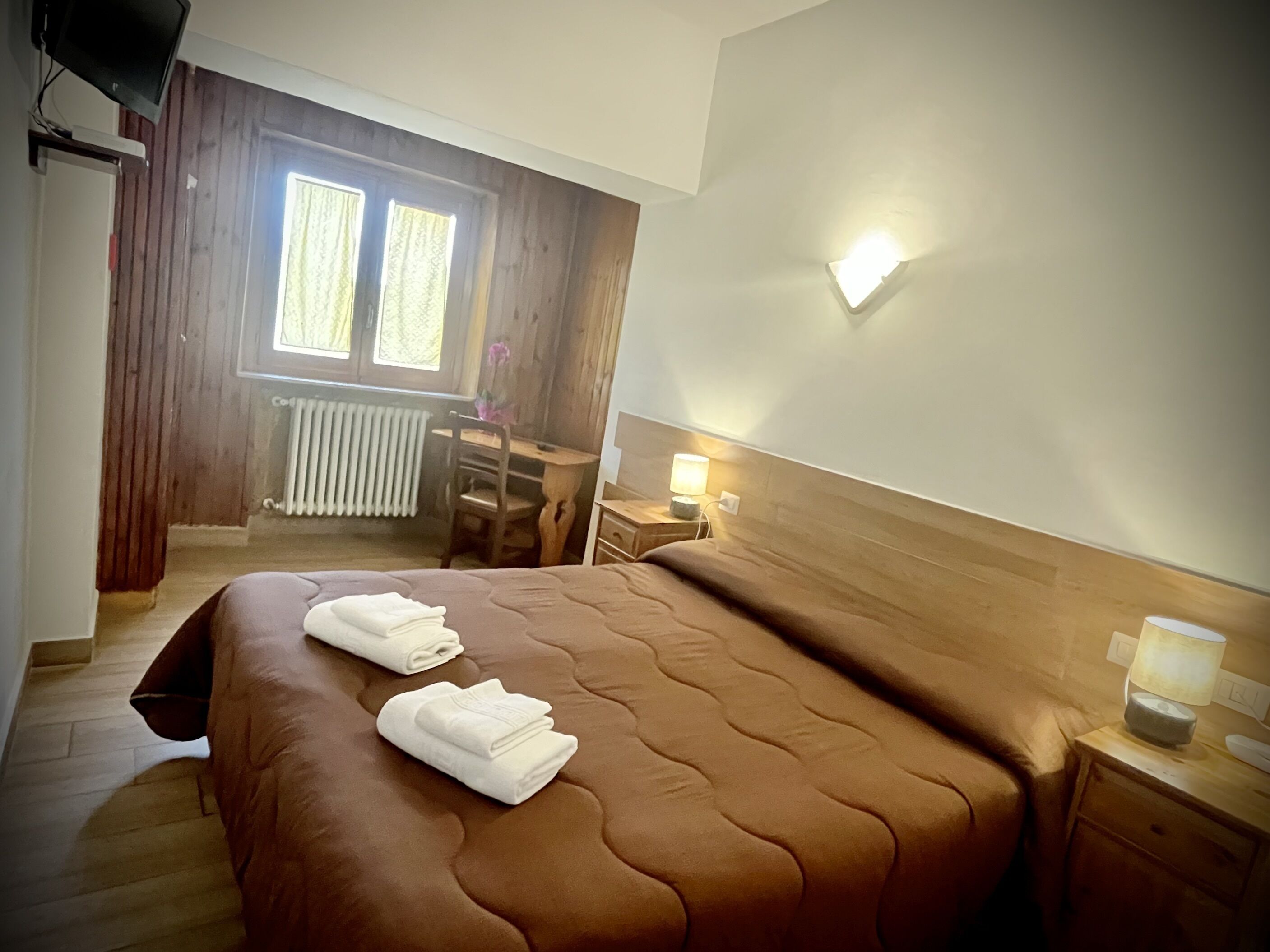 A hotel room with a large bed, a desk, a chair, a nightstand, a lamp, and a window with curtains.