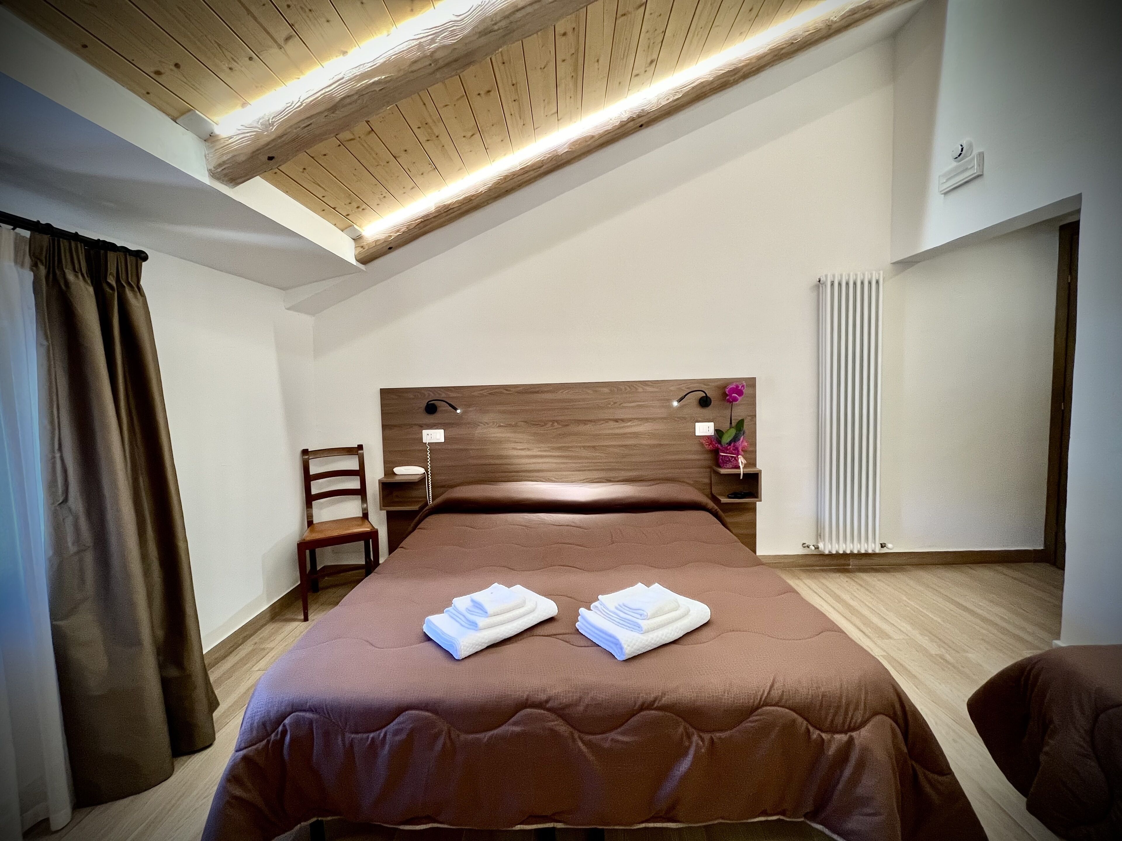 Ein Schlafzimmer mit einem hölzernen Kopfteil, einem Bett mit braunen Bettbezügen, einem Nachttisch mit Lampe, einem Sessel und einem Heizkörper.