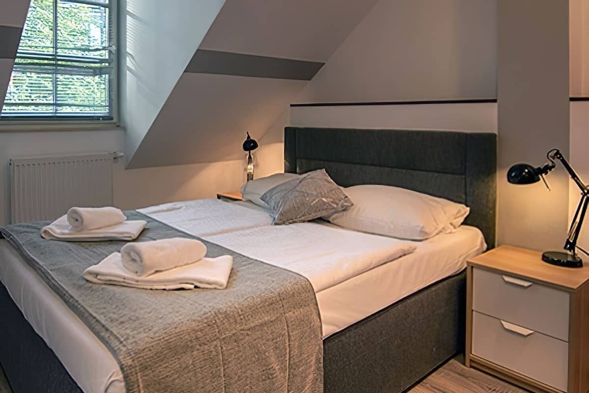 Una cama bien hecha con cabecera gris, una lámpara de noche y una mesita de noche con un cajón.
