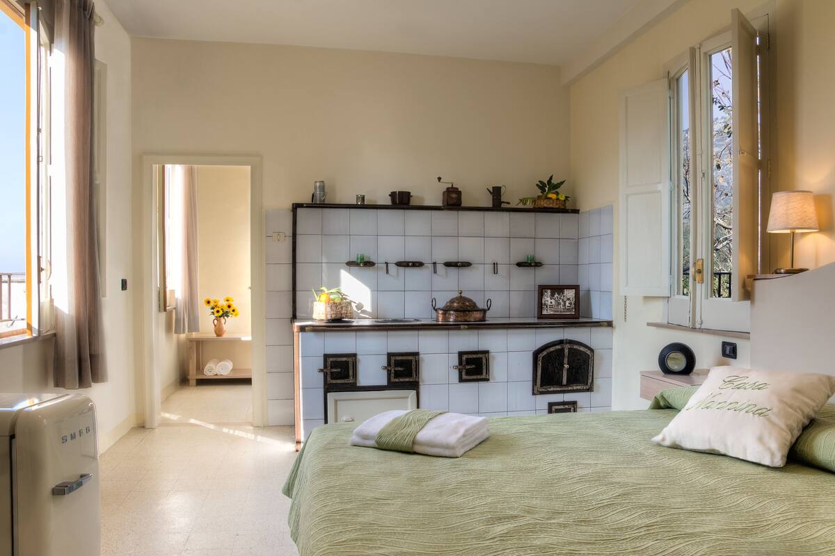 A bedroom with a bed, a nightstand, a window with curtains, a dresser, and a wall-mounted shelf with various items.