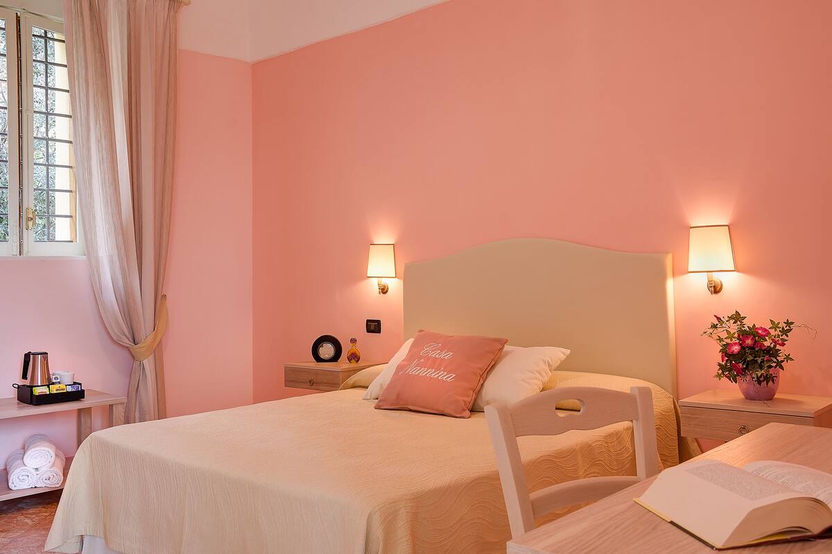 A neatly made bed with a pink pillow, bedside tables with lamps, a window with curtains, and a shelf with towels and a coffee maker.