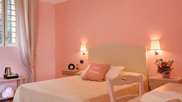 Cama bem arrumada com travesseiro rosa, mesas de cabeceira com luminárias, janela com cortinas e prateleira com toalhas e cafeteira.