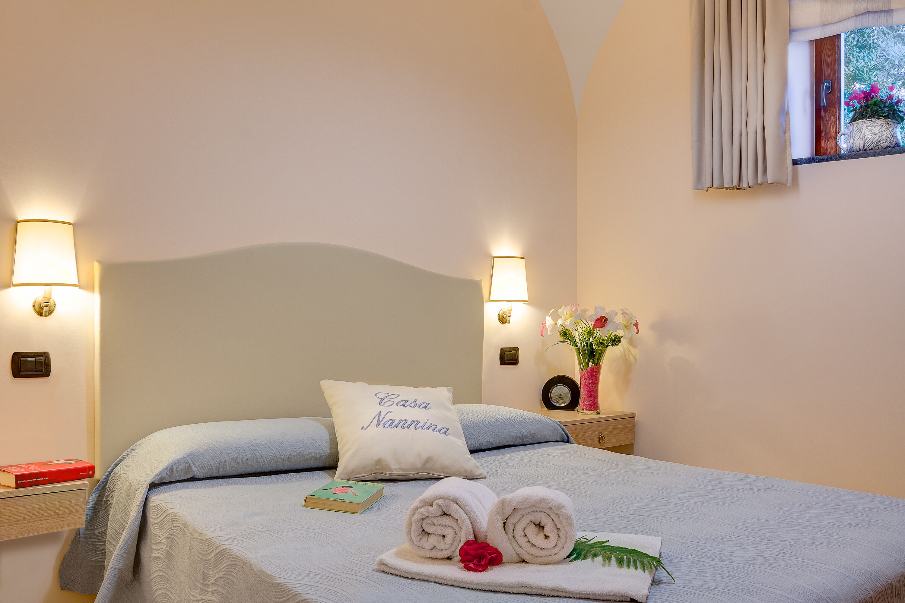 Una cama bien hecha con una almohada que dice "Casa Nannina", una mesita de noche con un libro y un pequeño jarrón de flores, y una ventana con cortinas.