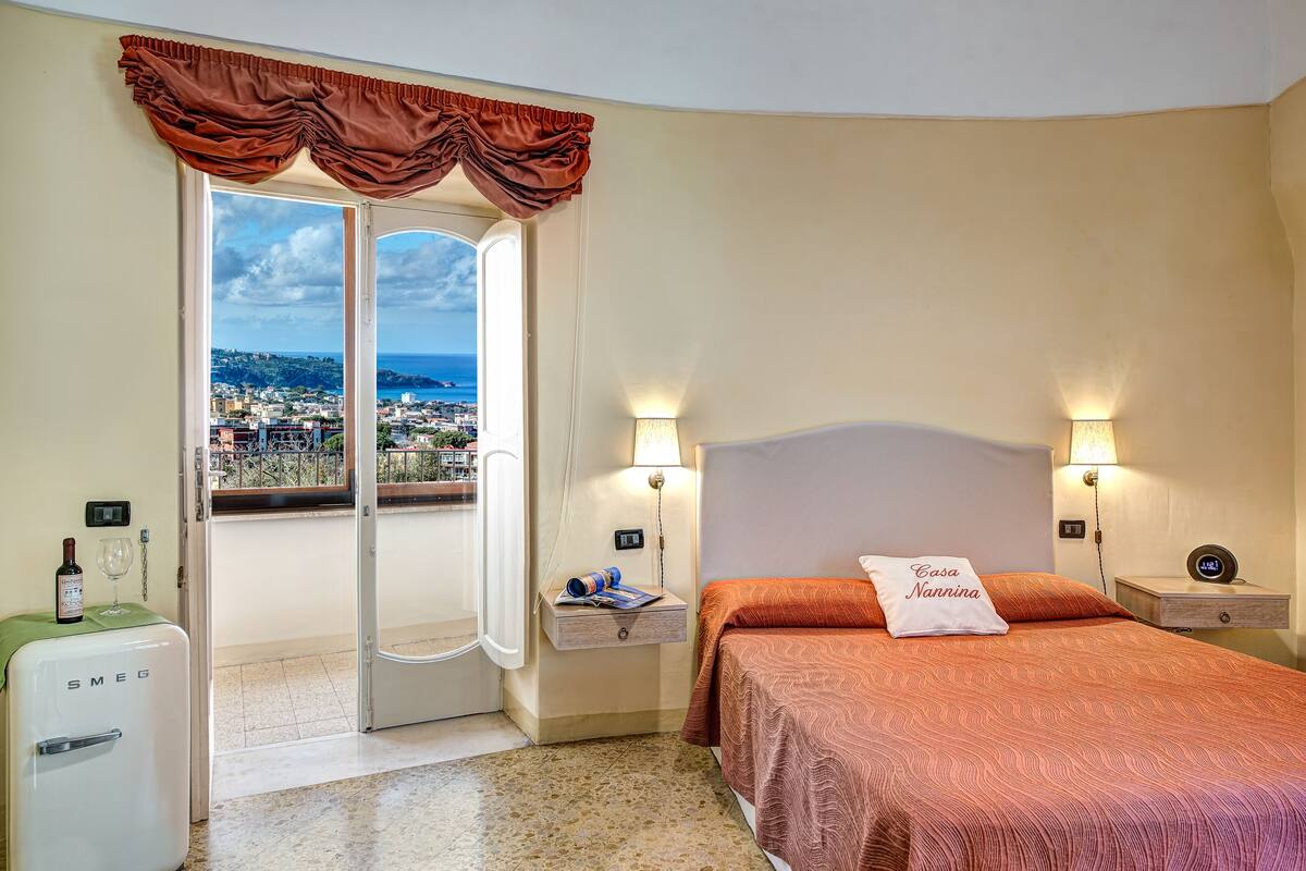 A hotel room with a bed, a nightstand, a lamp, a minibar, and a view of the city through a door.