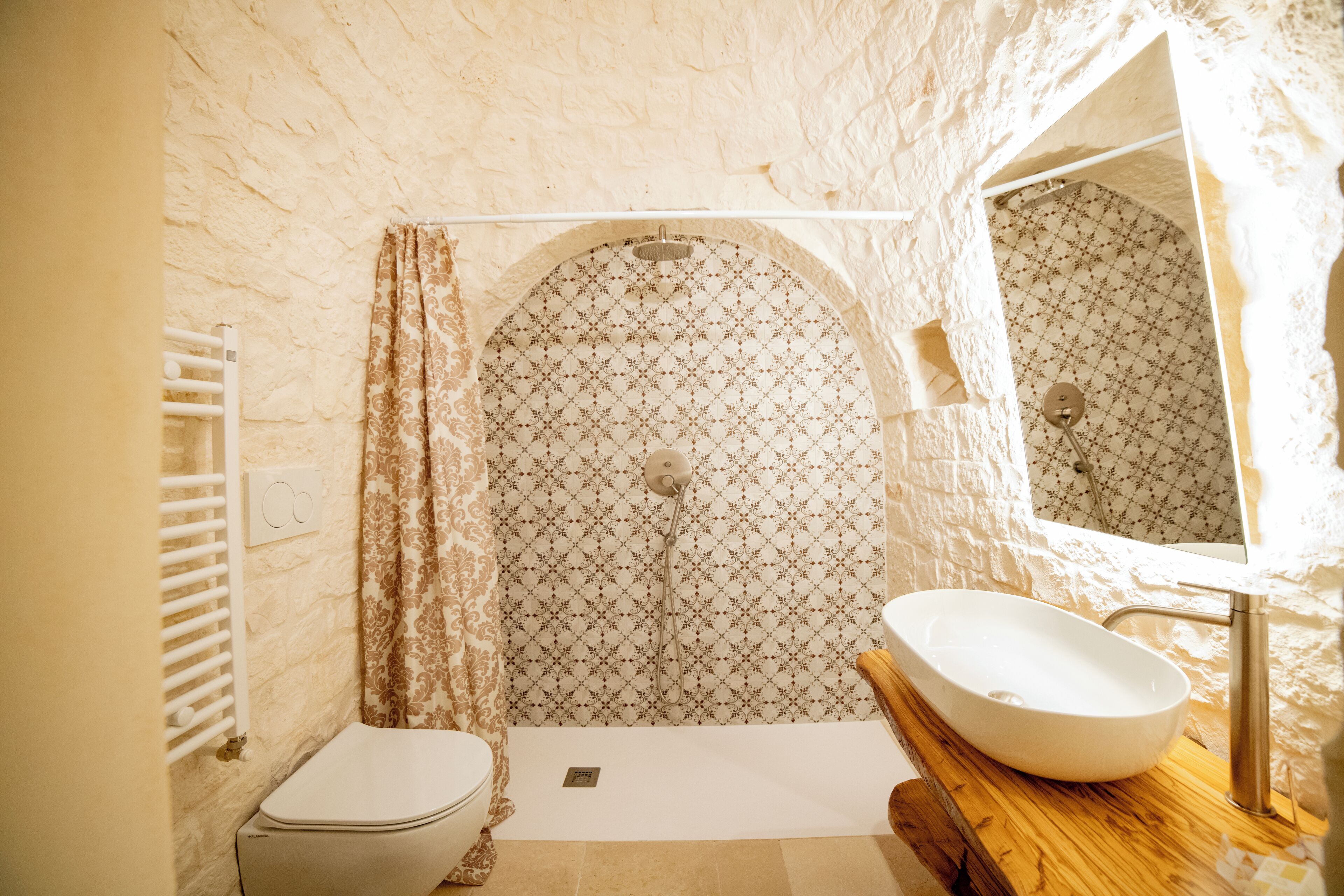 Une salle de bain avec des toilettes blanches, une douche avec un mur décoré et un meuble-lavabo en bois avec un évier.