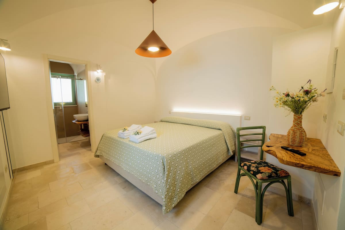 A bedroom with a bed, a chair, a wooden side table, a vase with flowers, and a bathroom visible through an open door.
