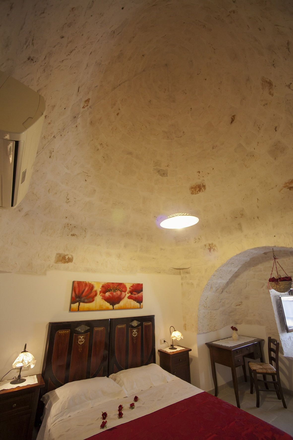 A bedroom with a wooden bed, a desk, and a chair. There is a painting on the wall and a ceiling light.