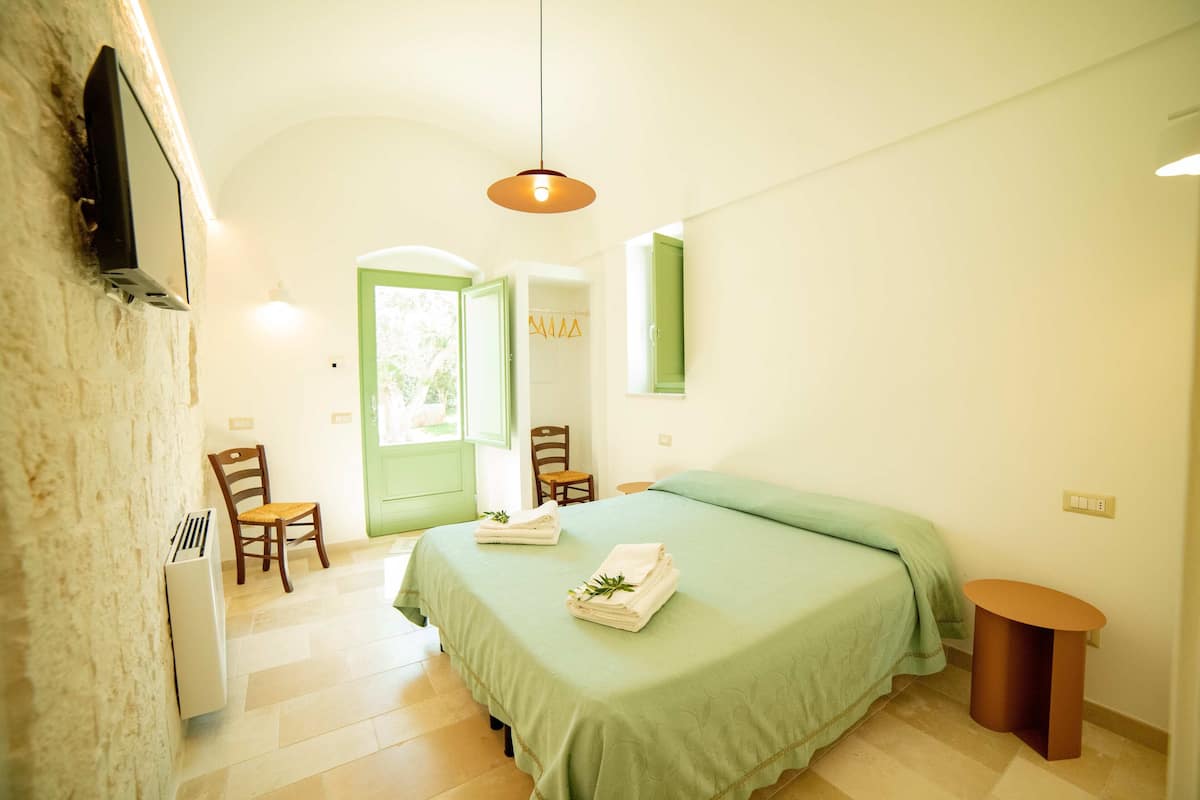A bedroom with a bed, two chairs, a small table, a wall-mounted TV, and a door leading to another room.