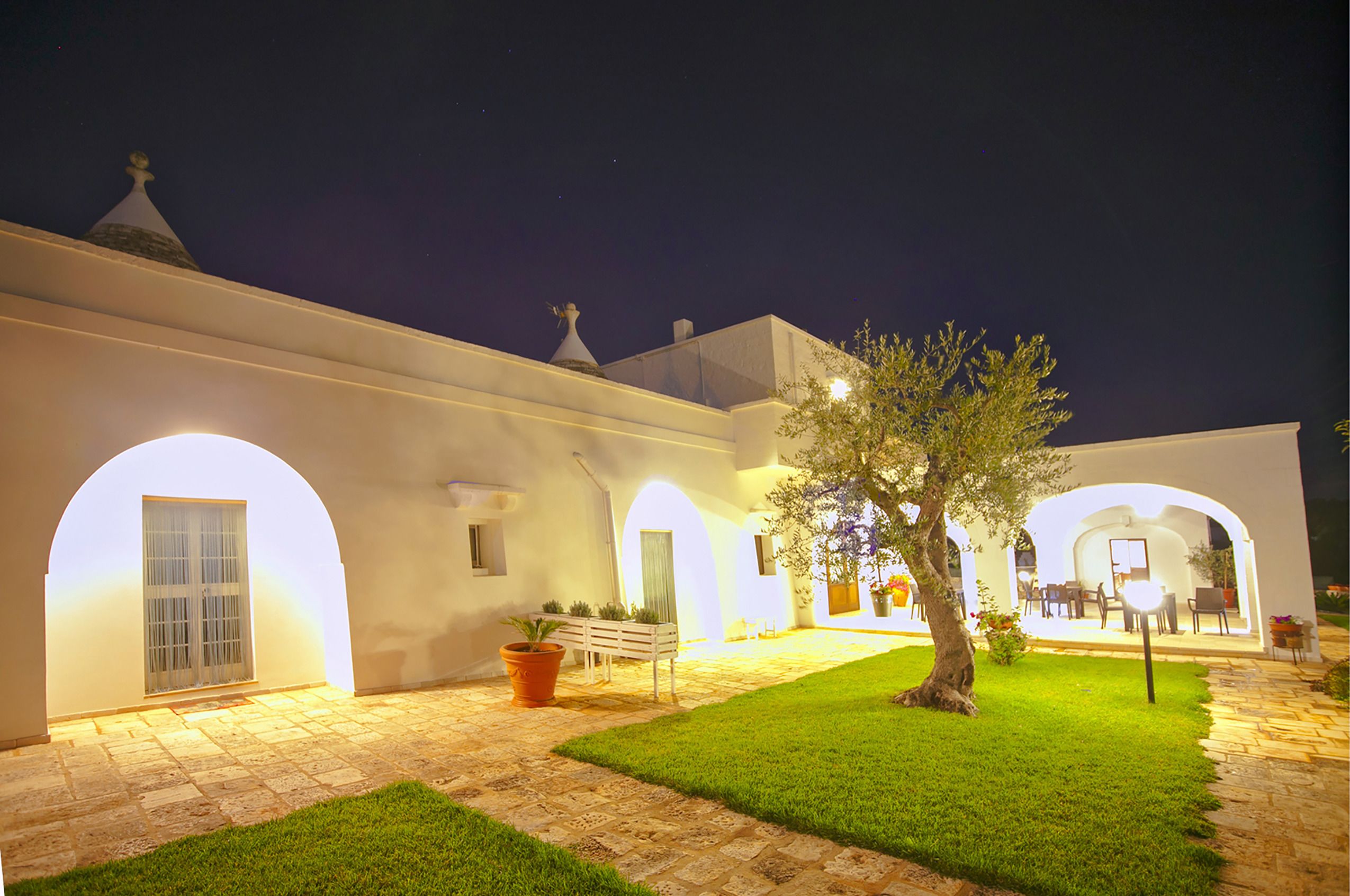 Un patio con un edificio blanco, arcos, un banco y un árbol.