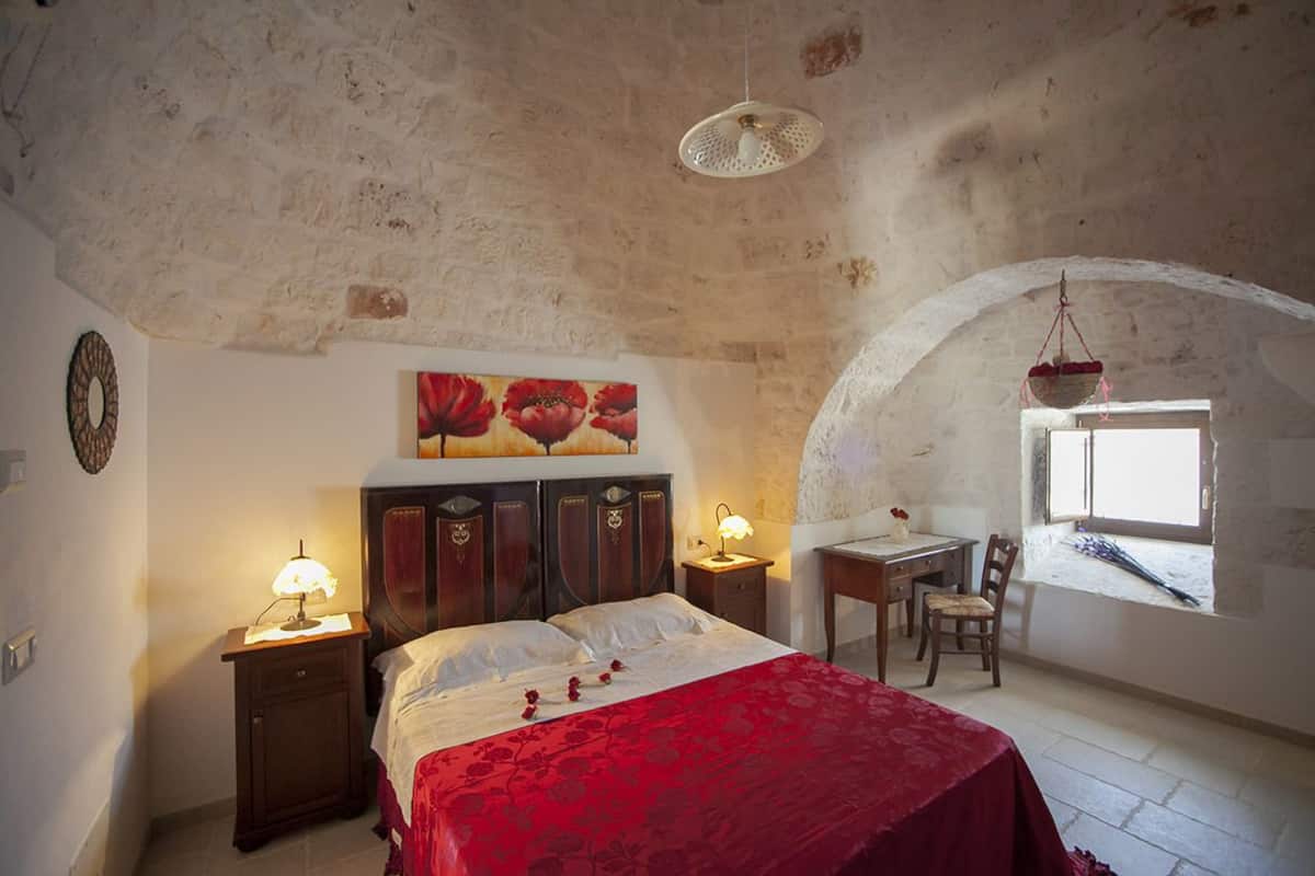 A bedroom with a large bed, two bedside tables with lamps, a wooden desk, and a chair. There is a window with a view of the outside.