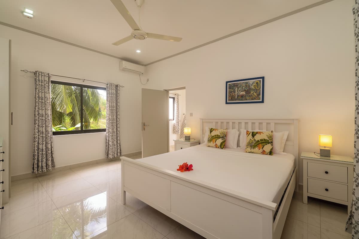 A bedroom with a bed, a ceiling fan, a nightstand with a lamp, and a window with curtains.