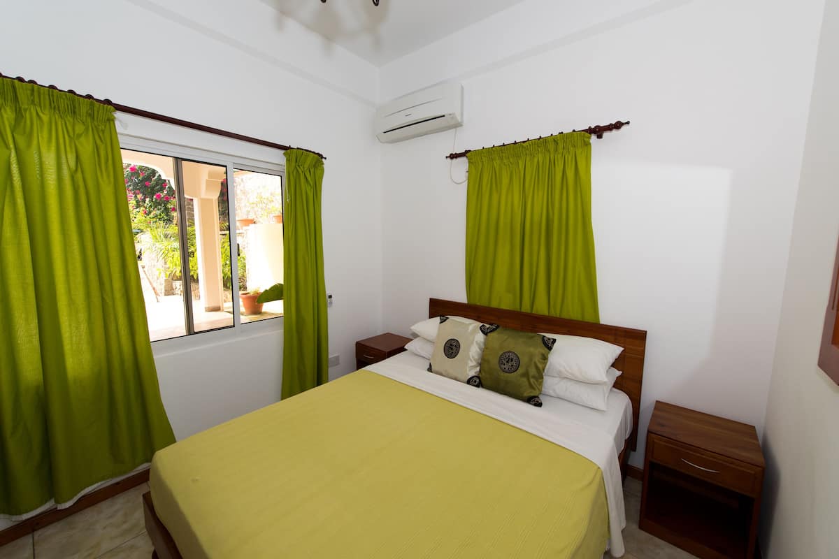 A bedroom with a bed, green curtains, a wooden nightstand, and an air conditioning unit.