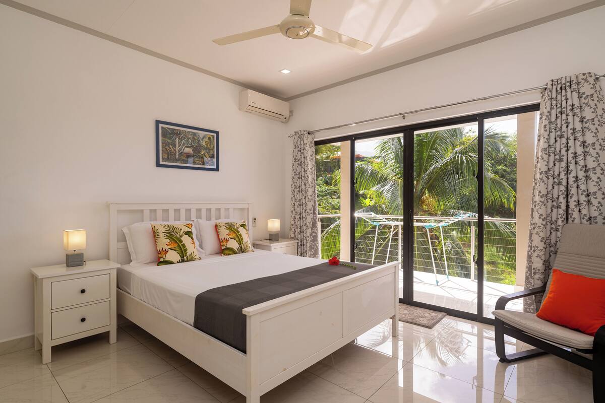 A bedroom with a bed, nightstands, a chair, a ceiling fan, and a large sliding glass door leading to a balcony with a view of greenery.