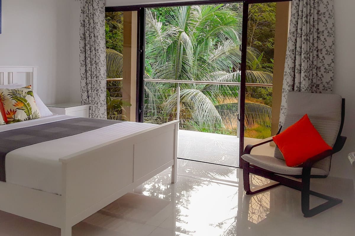 A bedroom with a bed, a chair, and a view of greenery through a glass door.