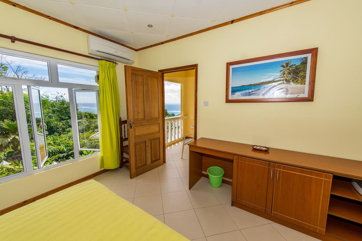 A room with a large window, a wooden desk, a green basket, and a framed picture of a beach.