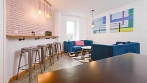Una sala moderna con un sofá azul, una mesa de centro de madera y una pared de ladrillo decorativa. Hay taburetes y un ventanal que proporciona luz natural.