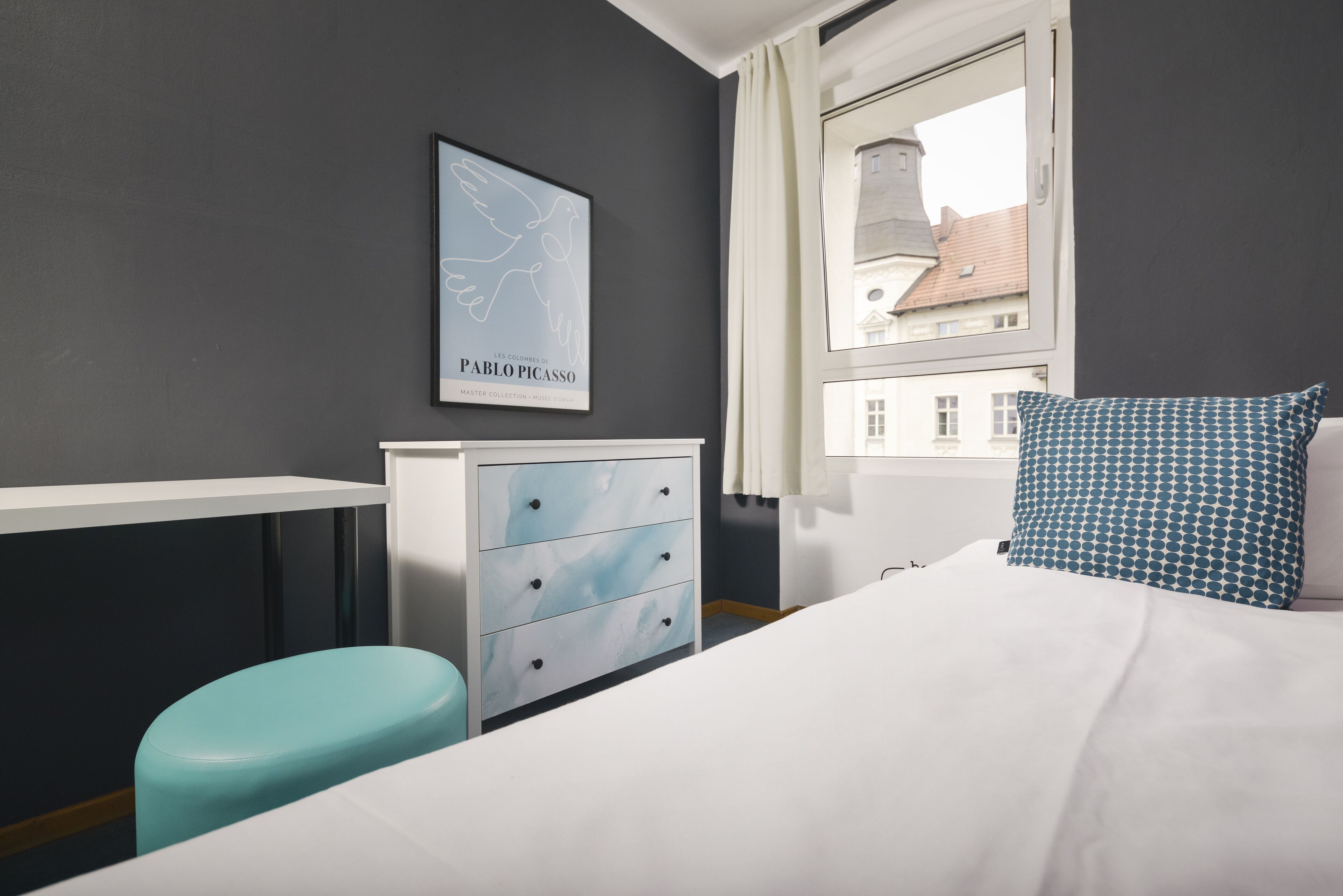 Ein Hotelzimmer mit einem Bett, einem blau-weißen Kissen, einer weißen Kommode mit Schubladen, einem gerahmten Kunstwerk an der Wand und einem Fenster mit Blick auf Gebäude.