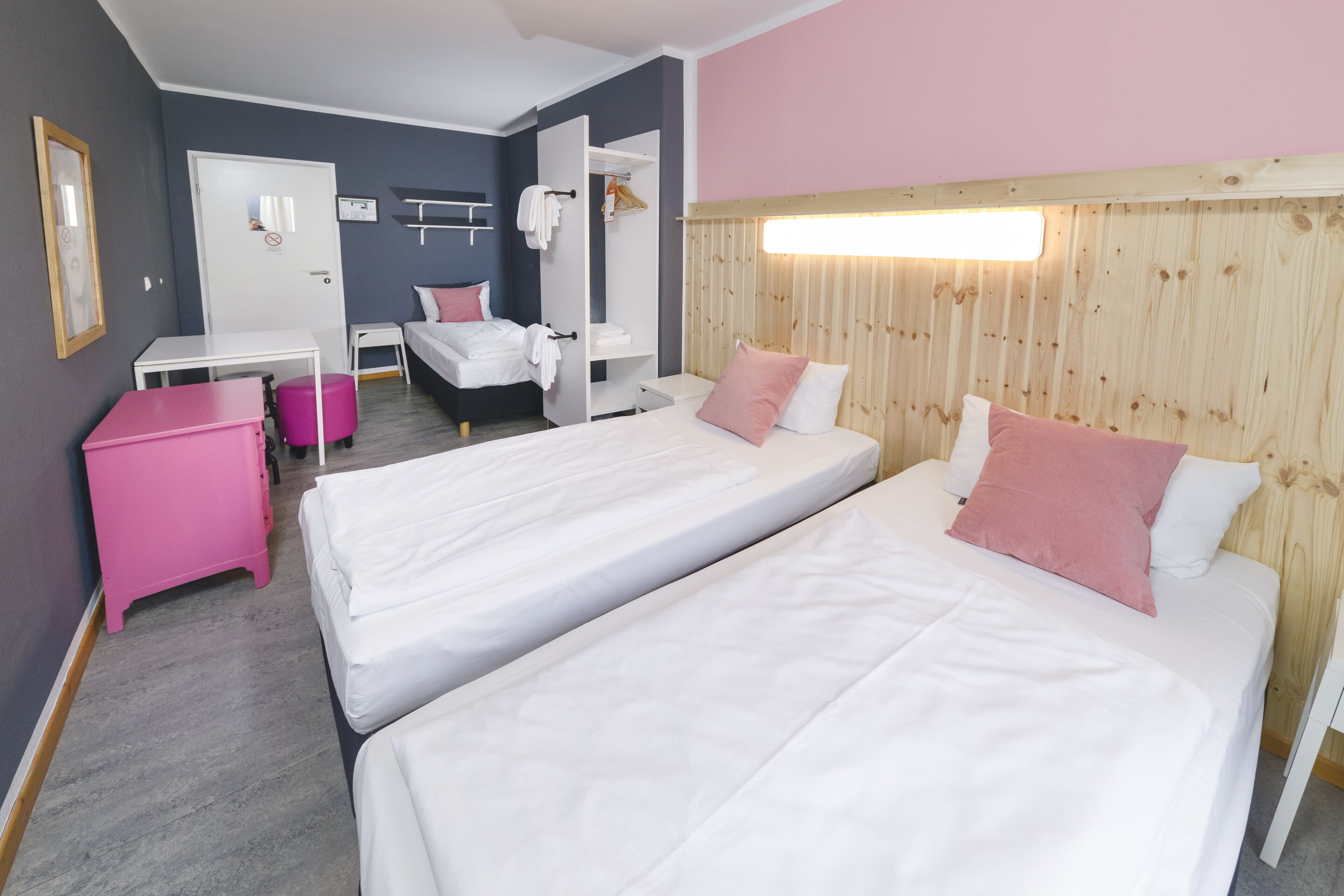 A hotel room with two beds, a pink dresser, a small table, and a wardrobe.